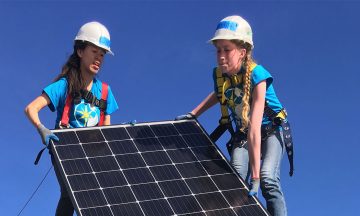 two students on top of a roof lift a solar panel up for installation
