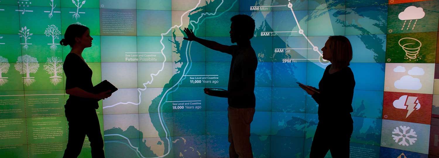 3 students stand in front of a large digital map of North America