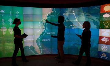 3 students stand in front of a large digital map of North America