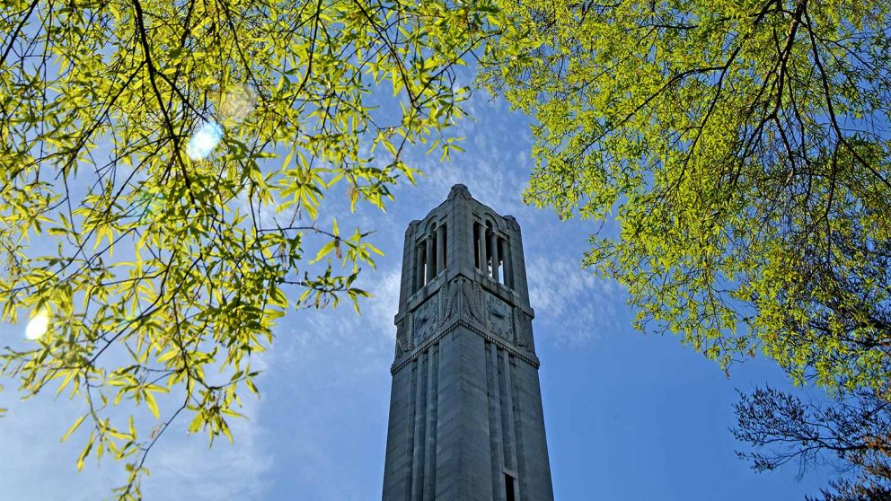 NC State Memorial Bell Tower is framed by green leaves with blue sky.