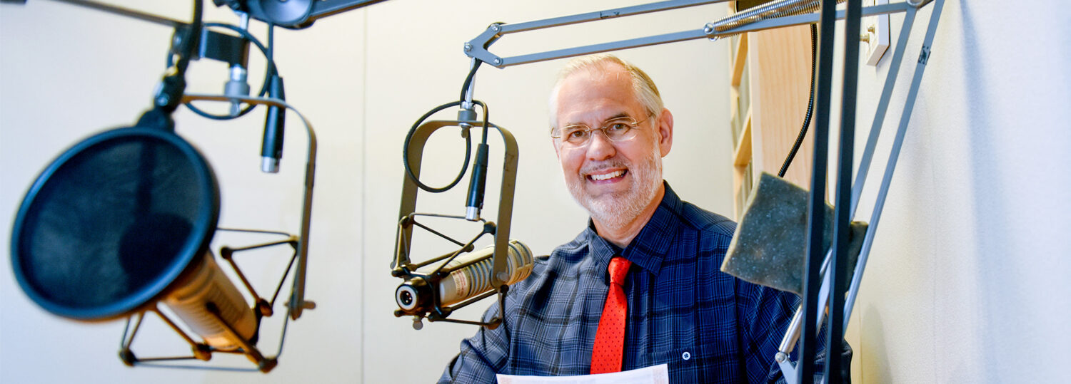 Walden sits in a recording studio with a red tie and dark blue plaid shirt.
