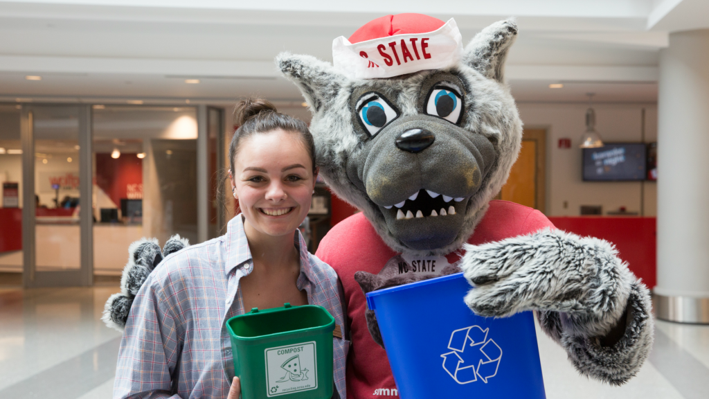 Student holds a mini green compost bin with Mr Wuf who is holding a blue recycling bin