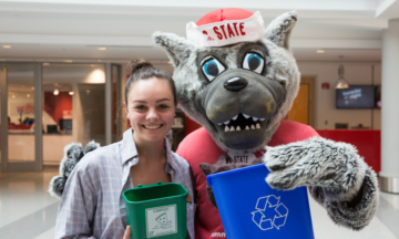 Student holds a mini green compost bin with Mr Wuf who is holding a blue recycling bin