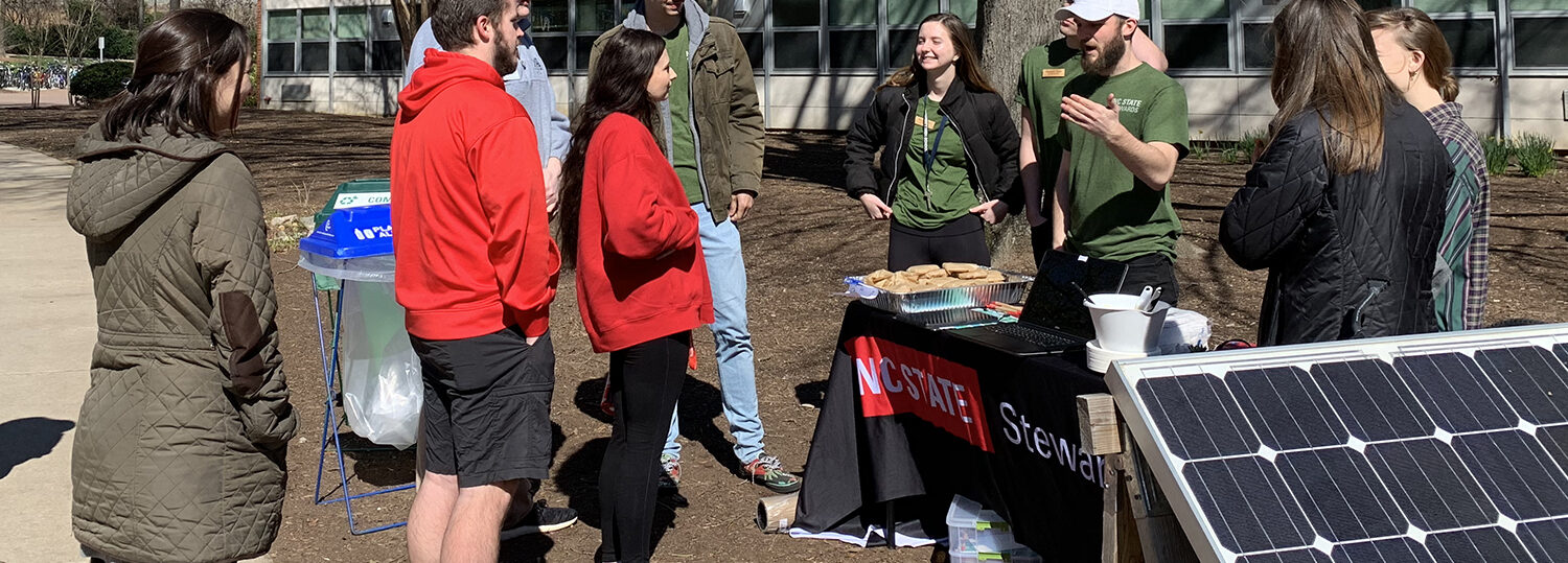 NC State Stewards, wearing matching green T-shirts, speak to a small group of fellow students outside next to a solar panel.