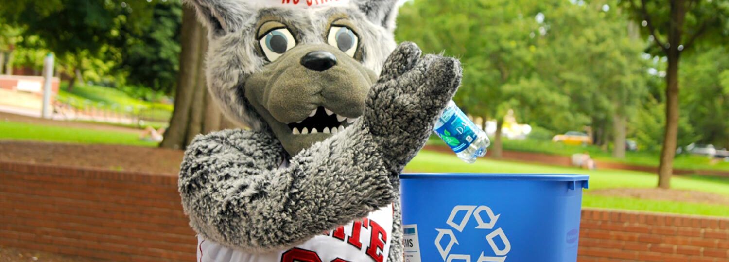 Mr Wuf holding a small blue recycling bin and putting a plastic water bottle inside while standing outside on campus grounds