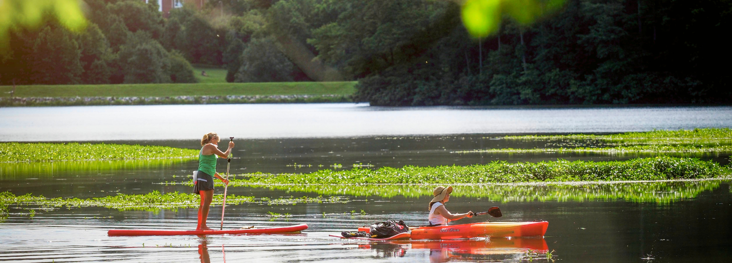 A kayaker and a stand up paddler enjoy Lake Raleigh with views of Centennial Campus behind them.