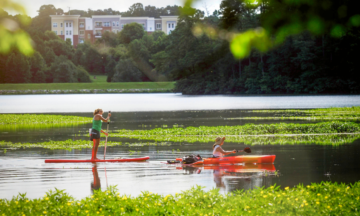 A kayaker and a stand up paddler enjoy Lake Raleigh with views of Centennial Campus behind them.