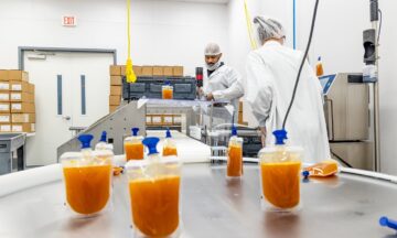 Two people in white lab coats and hair nets work at machinery. Packets of orange puree are in the foreground.