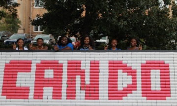A group of students pose behind a wall of canned goods organized in a way that spells out "Can Do" in red and white