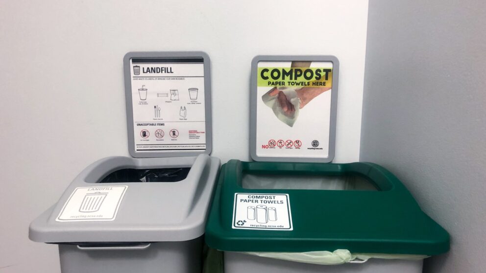 A photo of two bins placed next to each other in the corner of a Wilson College of Textiles bathroom. One is a waste bin and is gray. One (right) is a green bin and is for composting. Both have signage above them explaining what items to place in each type of bin.