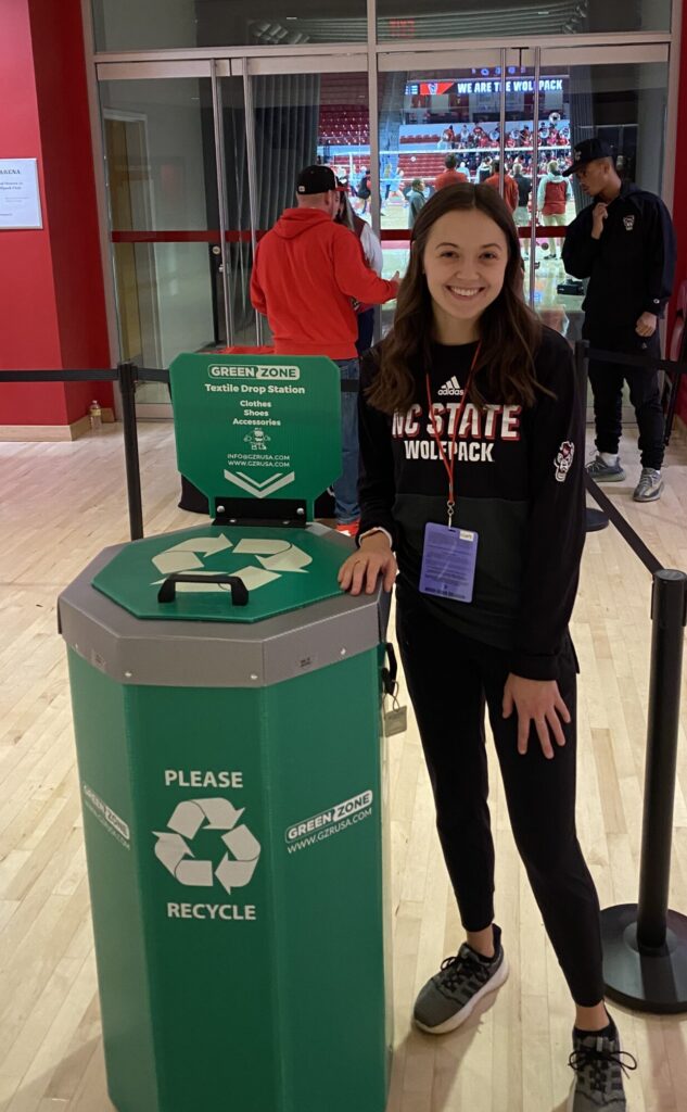 Claire Henson stands next to a textile recycling bin in the lobby of Reynold Gym while Volleyball game is being played.