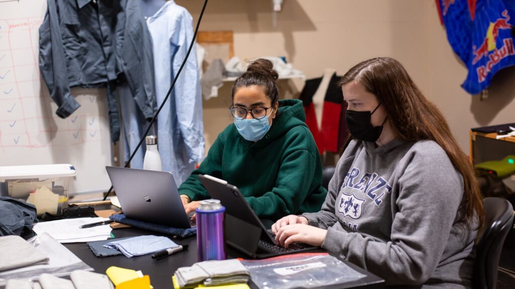 Two students work at a table in the Senior Design Lab. Textile samples are on the table they are working at, and the PPE suit they are working to improve is visible hanging on a whiteboard behind them.