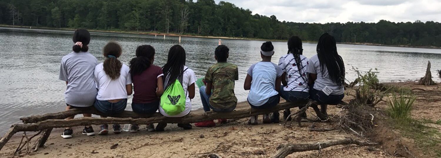 A group of middle school students sit on a log and look out onto a lake.
