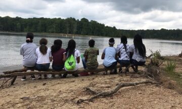 A group of middle school students sit on a log and look out onto a lake.