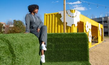 Delisia Matthews wears a long grey dress with with white drawstrings at the collar and long sleeves. She sits on top of a larger-than-life green couch made out of synthetic turf with her legs crossed and looking straight ahead but away from the camera, which is positioned to her right. Behind her, a remodeled shipping container that has been turned into a work space is visible.