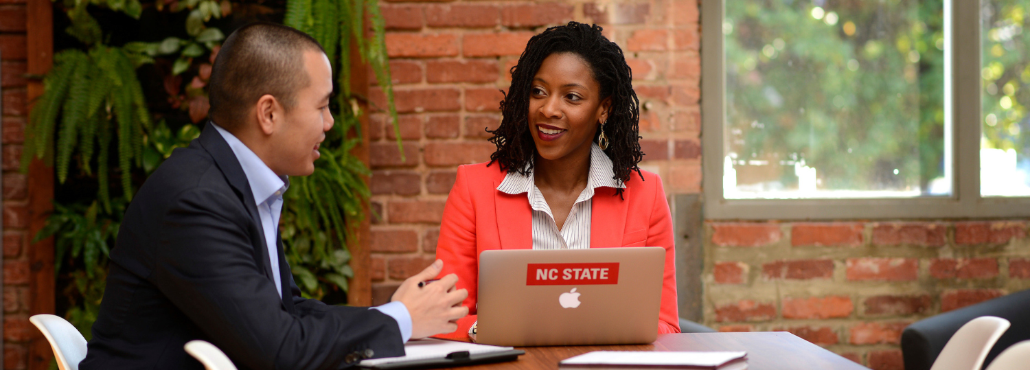 2 Poole College MBA students collaborate at a long wood table with an NC State laptop open and brick wall behind them. Photo by Marc Hall