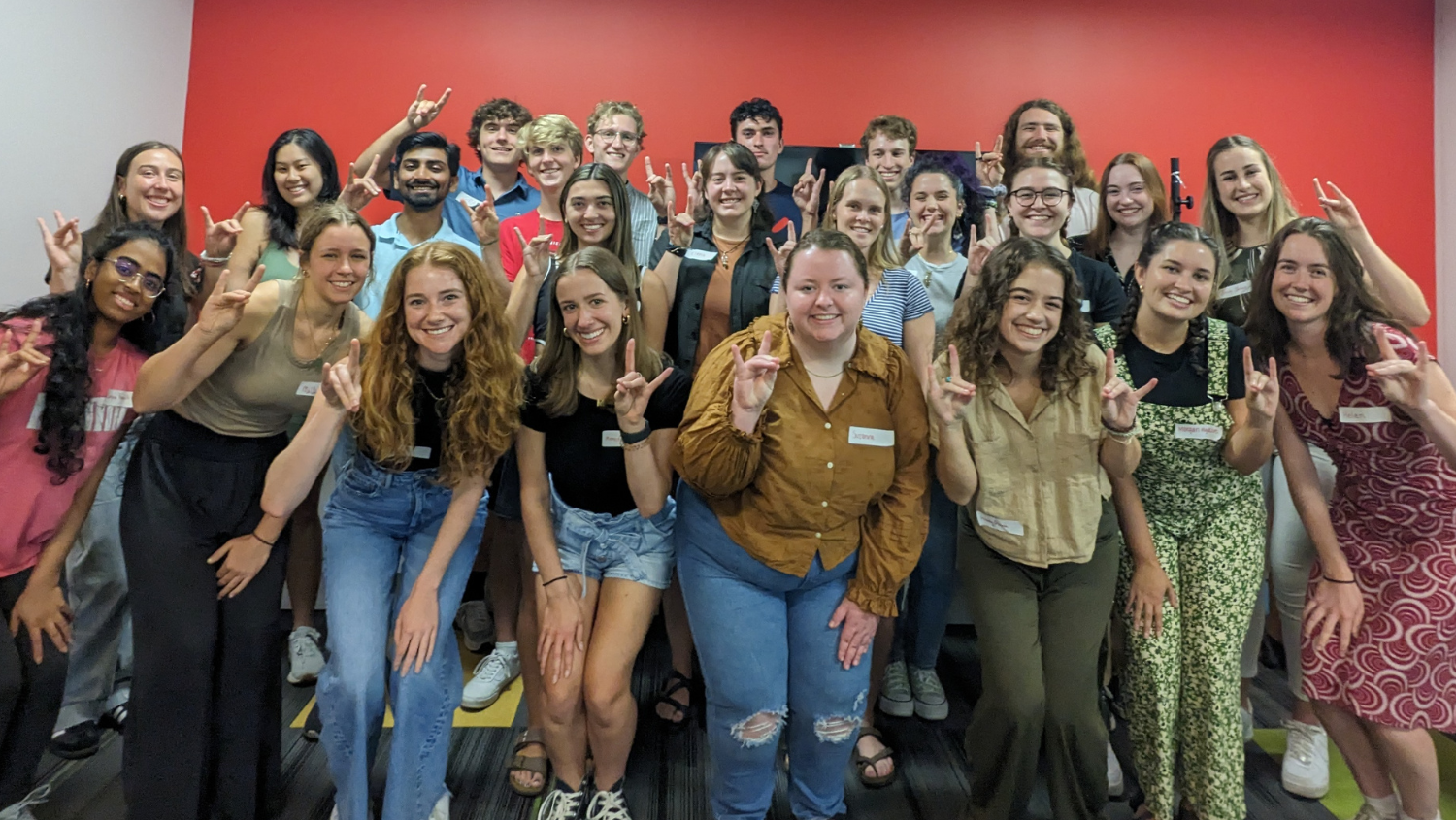 Campus As A Classroom Fall 2023 cohort photo, group  of students showing wolfies hand gesture in front of a red wall