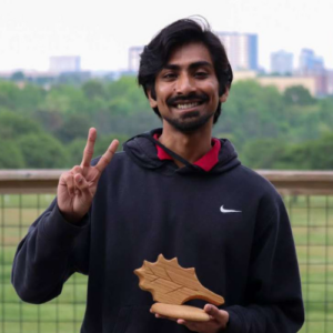 Kunnumpurath Jayakumar standing with City of Raleigh Environmental Award APril 2024 giving peace sign with Raleigh skyline in background