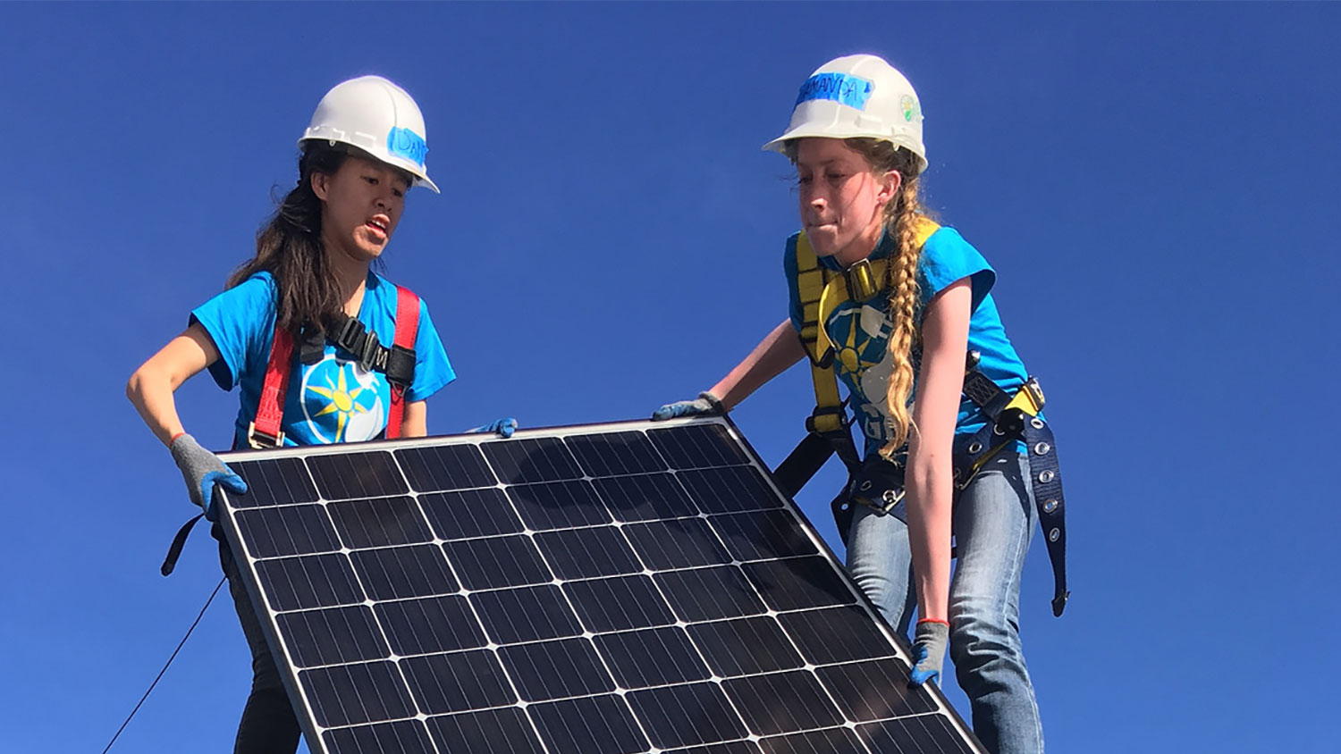 two students on top of a roof lift a solar panel up for installation