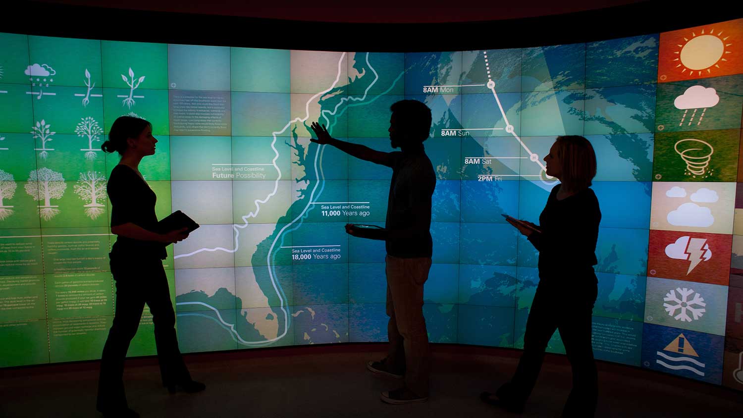 3 students stand in front of a large digital map of North America