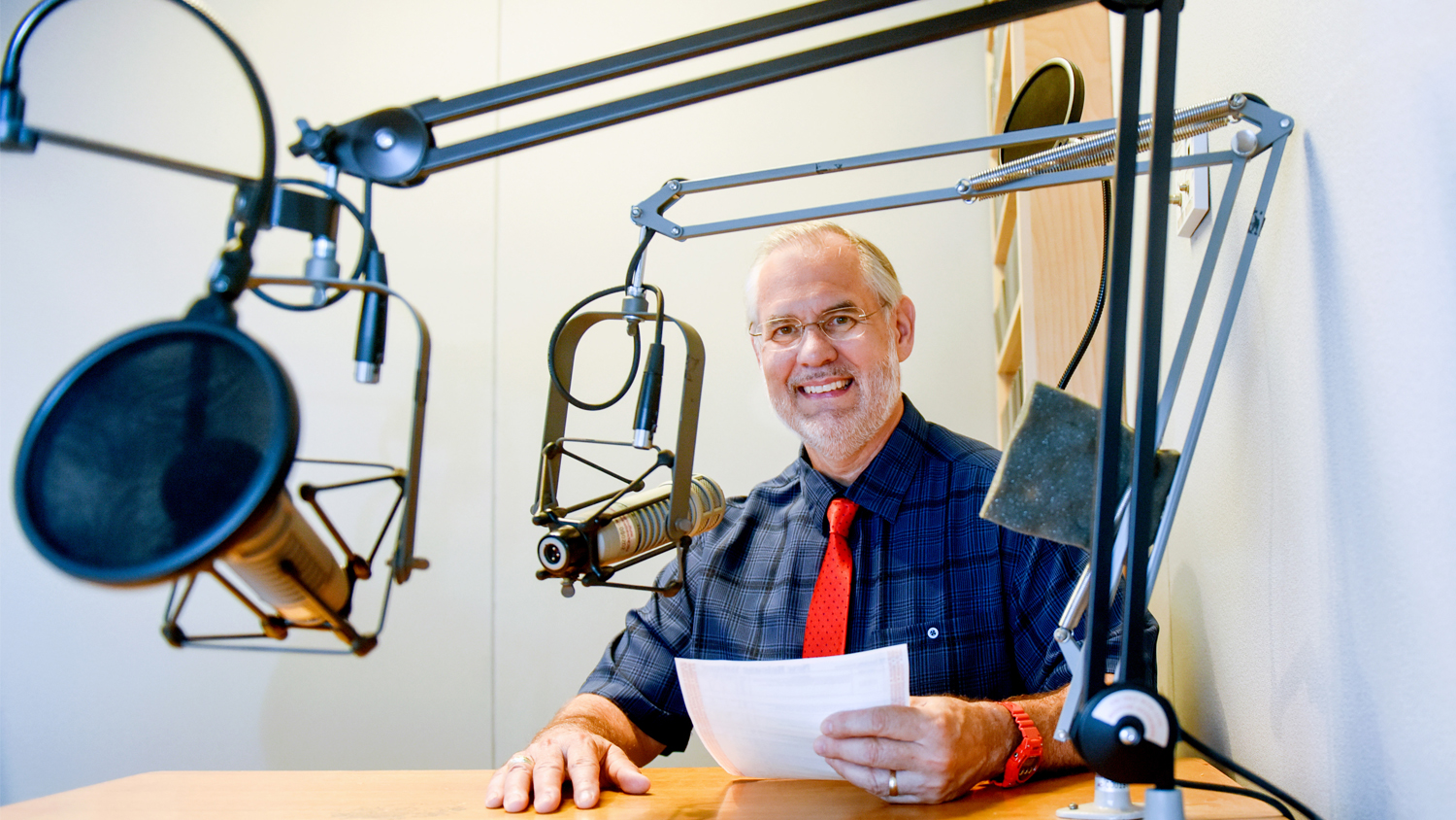 Walden sits in a recording studio with a red tie and dark blue plaid shirt.