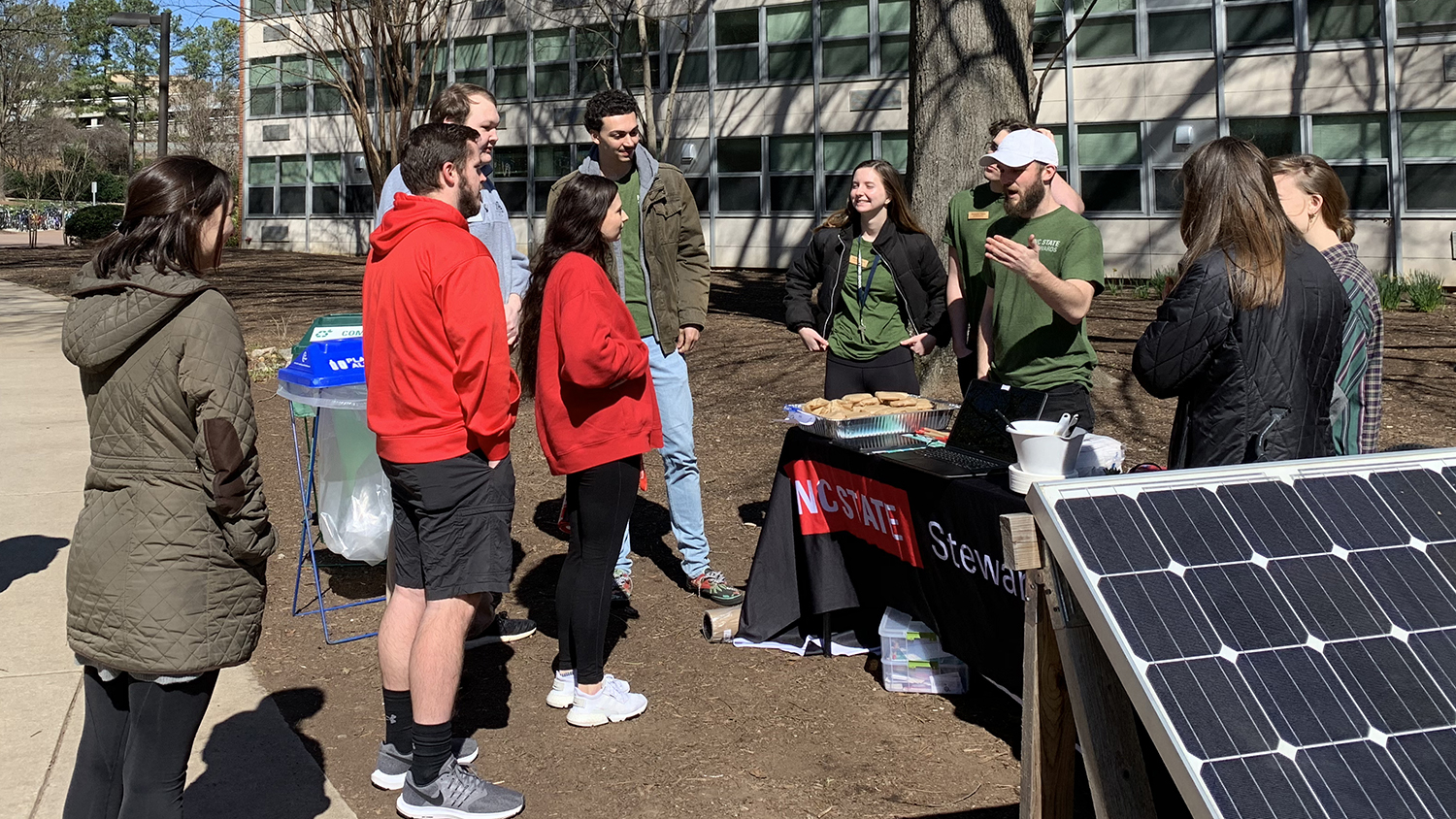 NC State Stewards, wearing matching green T-shirts, speak to a small group of fellow students outside next to a solar panel.