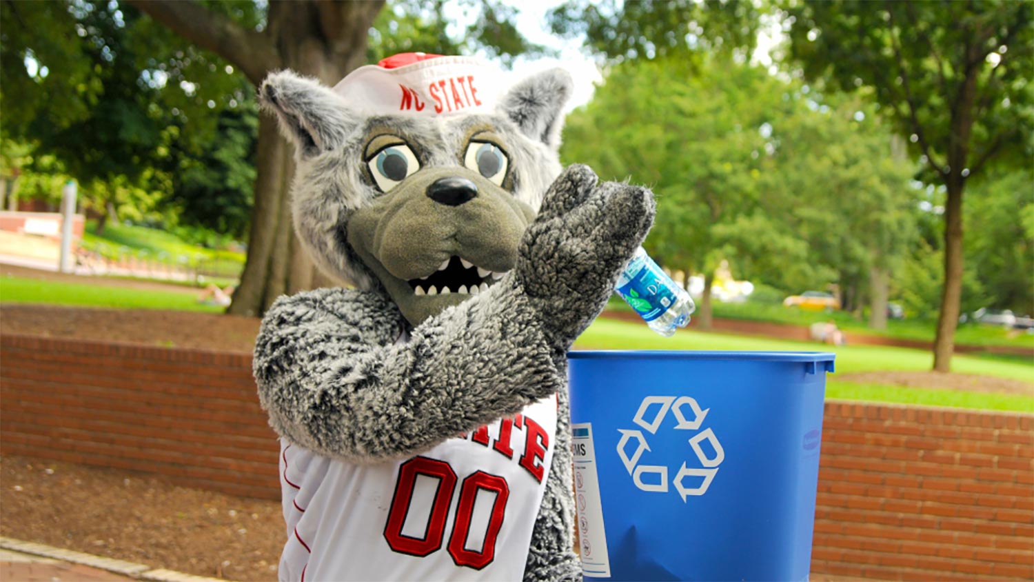 Mr Wuf holding a small blue recycling bin and putting a plastic water bottle inside while standing outside on campus grounds