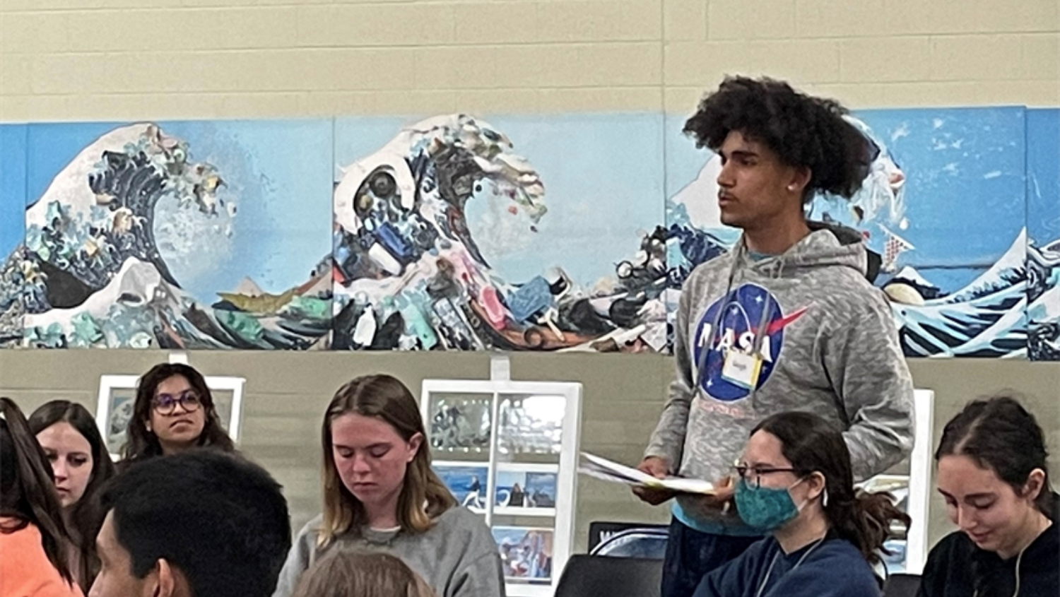 student wearing NASA hoodie stands talking in meeting in front of ocean mural