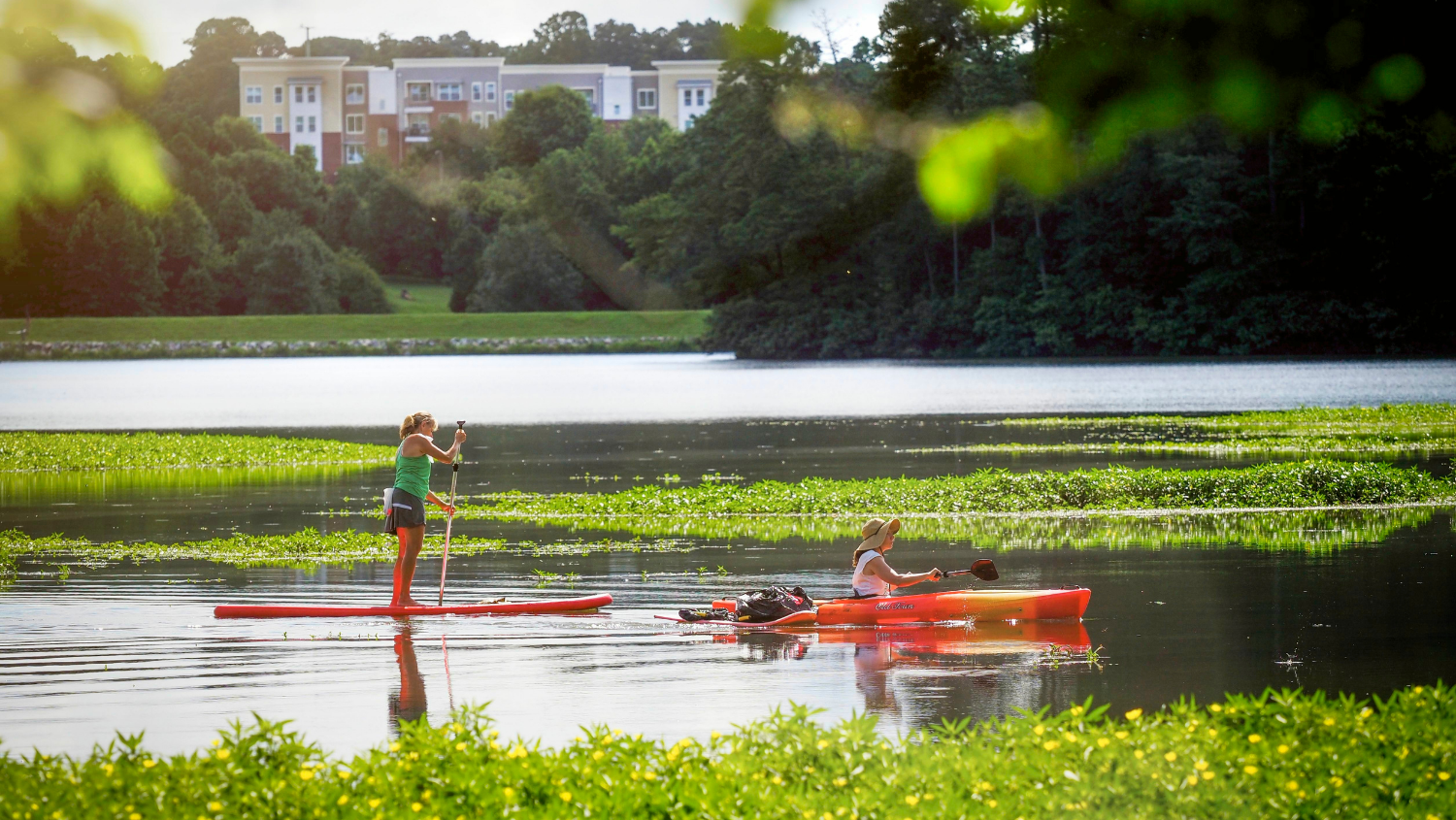 A kayaker and a stand up paddler enjoy Lake Raleigh with views of Centennial Campus behind them.