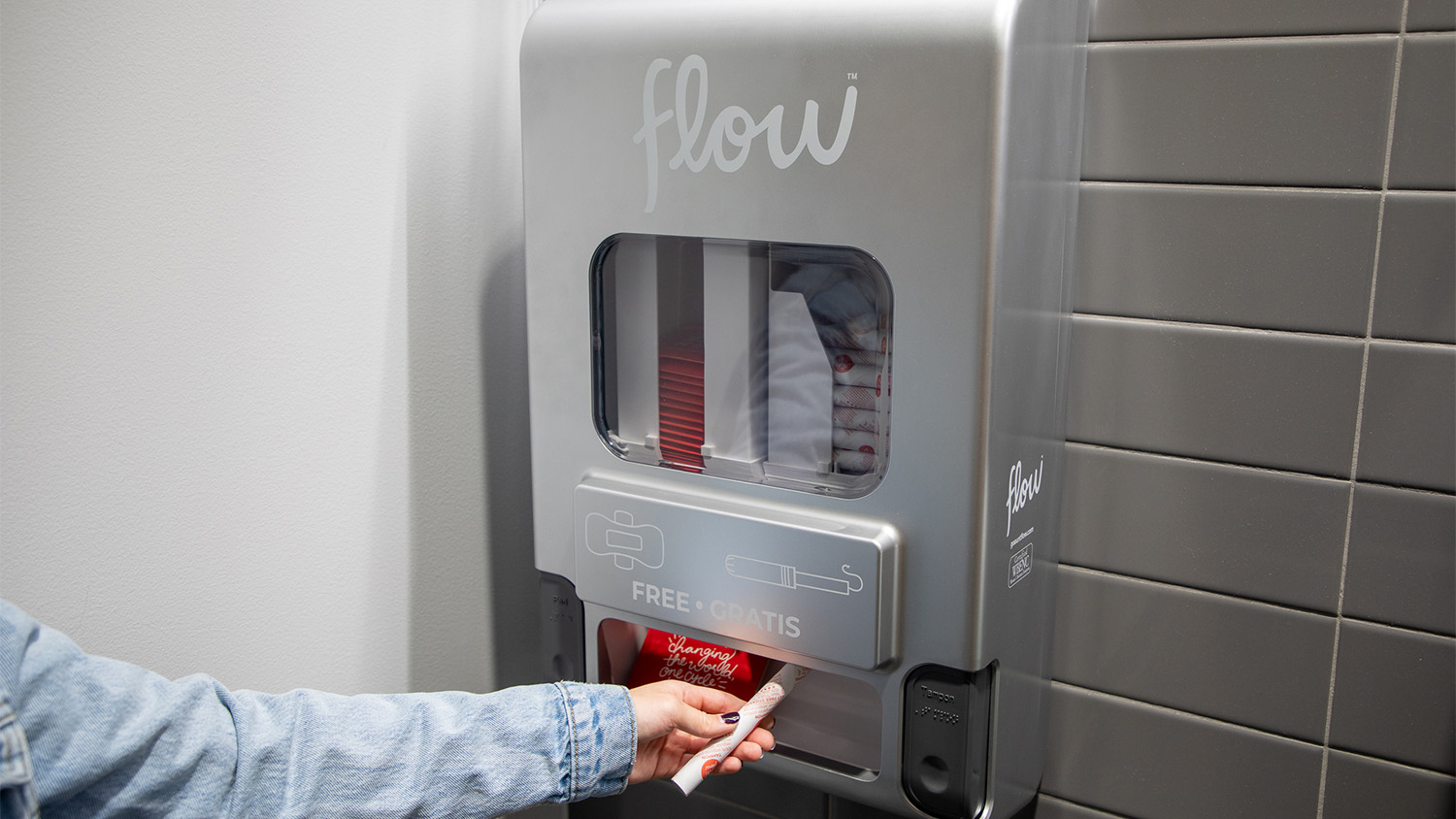 A hand pulls a tampon from a dispenser on a restroom wall