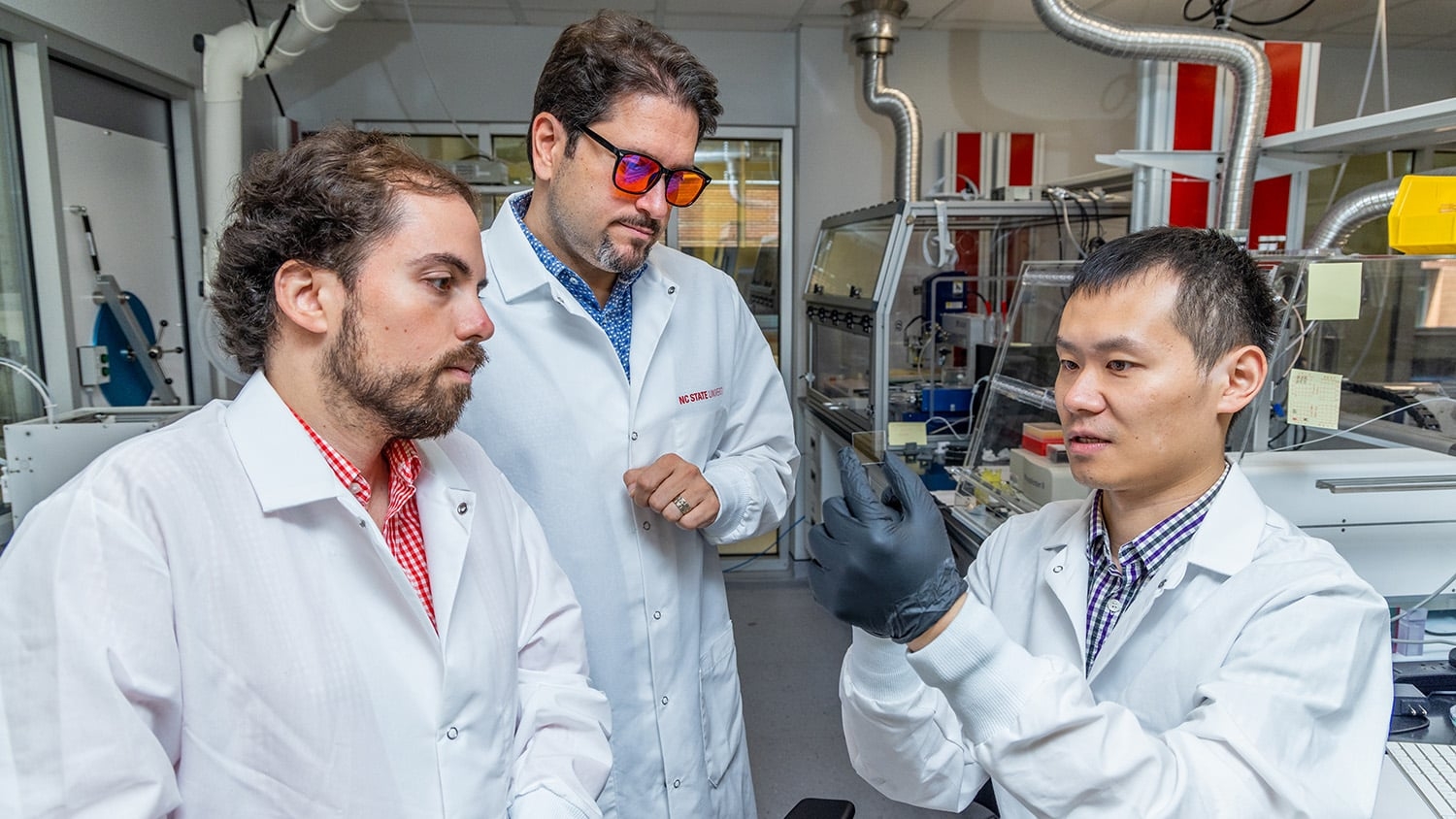 From left, Nathan Woodward, Aram Amassian and Tonhui Wang examine a microscope slide. The three wear white lab coats, and Wang, who is holding the slide, wears black gloves.