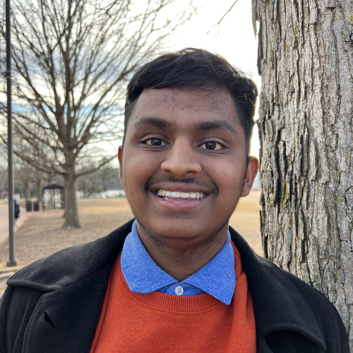 Skanda Vasuki Steward outside in blue collar shirt over orange sweater with black jacket