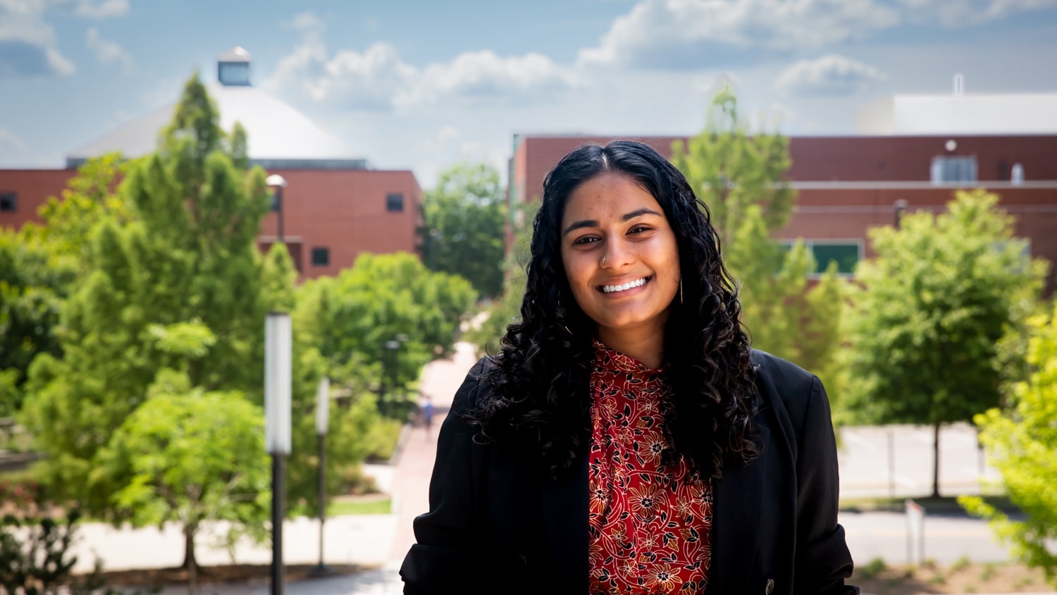 Jyotsna Gopinath stands on the stairs next to Hunt Library with the Wilson College of Textiles and bright green trees visible in the background.