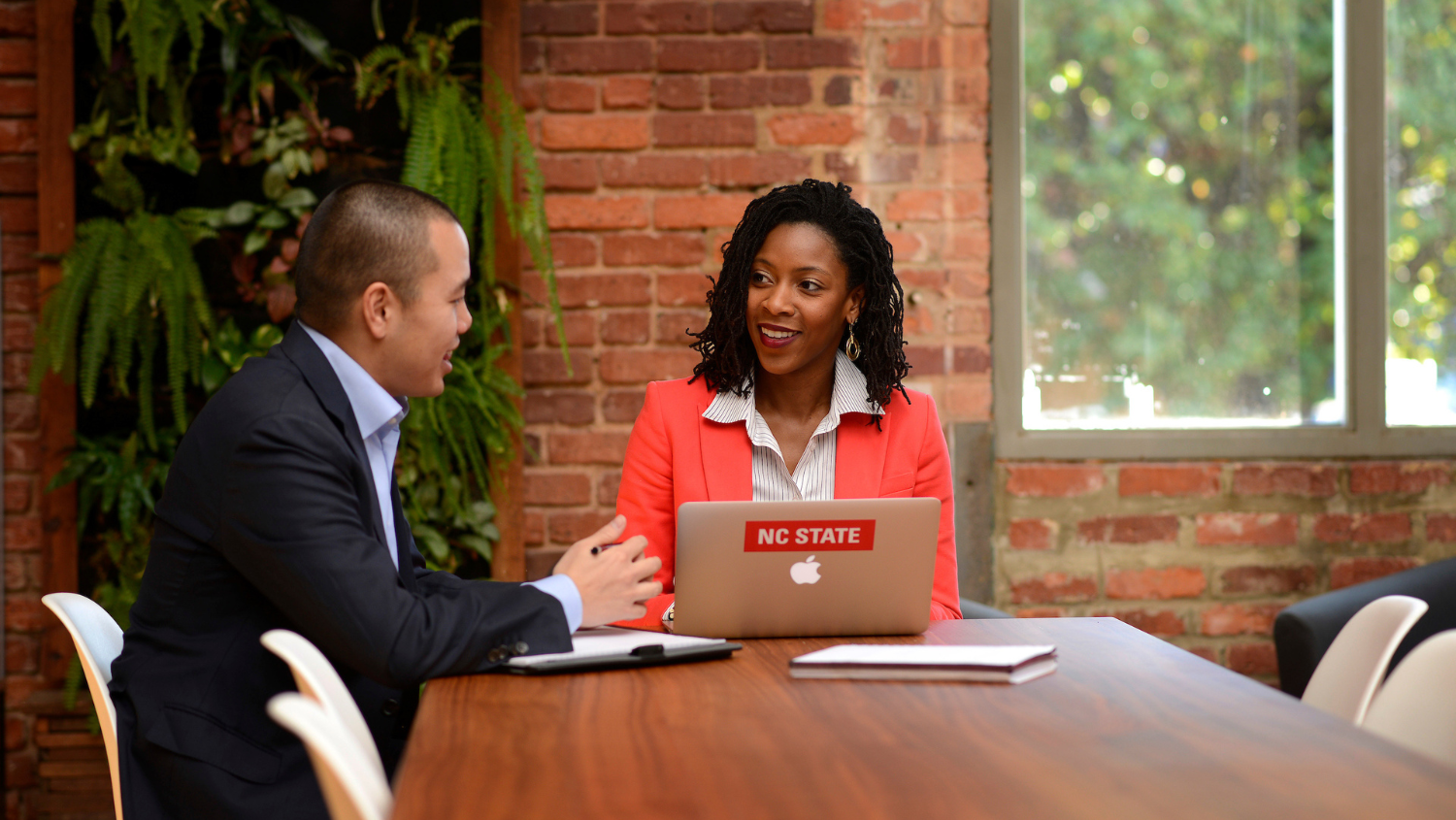 2 Poole College MBA students collaborate at a long wood table with an NC State laptop open and brick wall behind them. Photo by Marc Hall