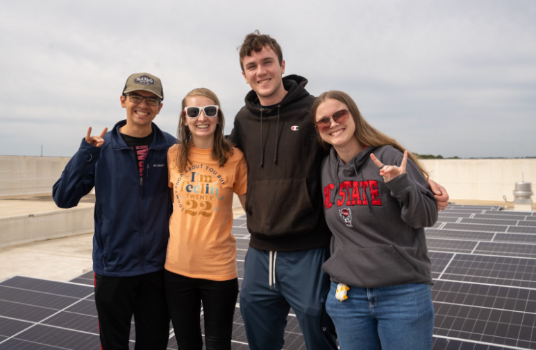 4 Students stand on top of Fitts-Woolard hall with solar panels in background. They are members of the ECE Senior Design Team