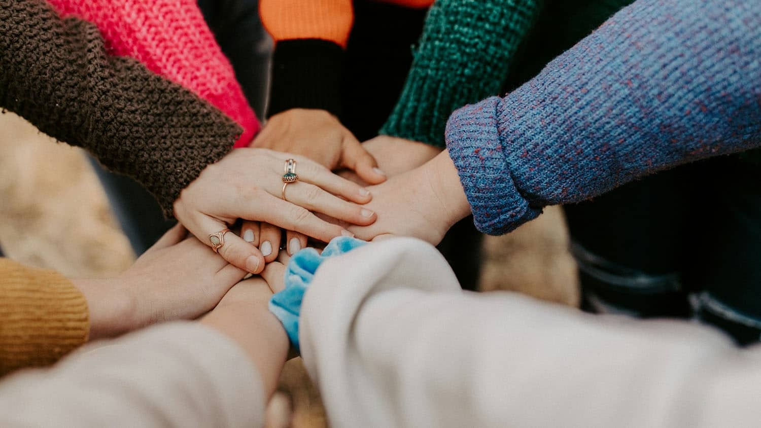 eight people standing in a circle have put one hand each into the center, symbolizing collective teamwork and effort